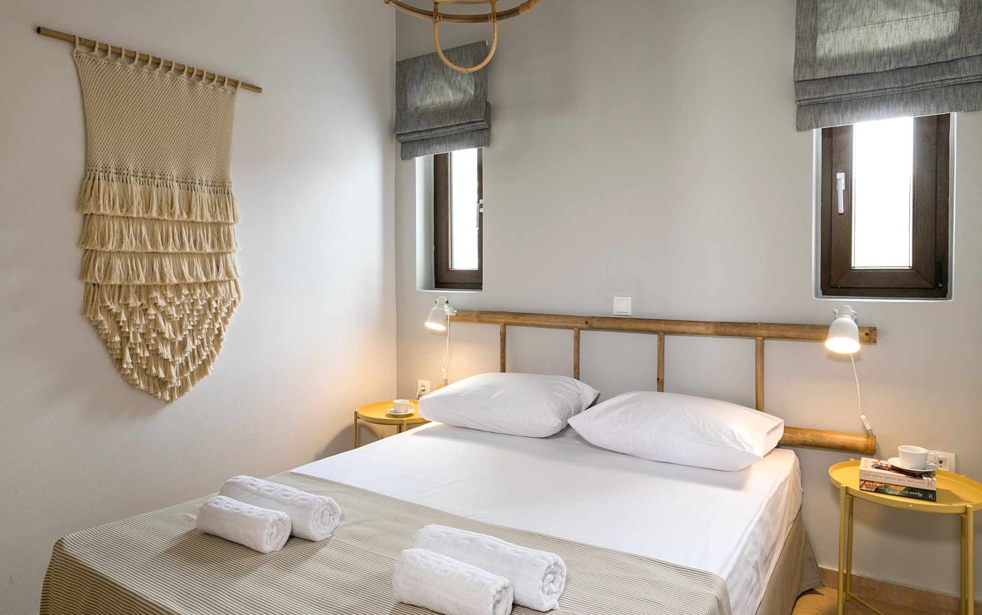 Syra Suite bedroom with a double bed, metallic yellow bedside tables, modern white wall lights, two windows on the wall over the bed and boho decoration with a wooden ladder over the bed and a big macramé.