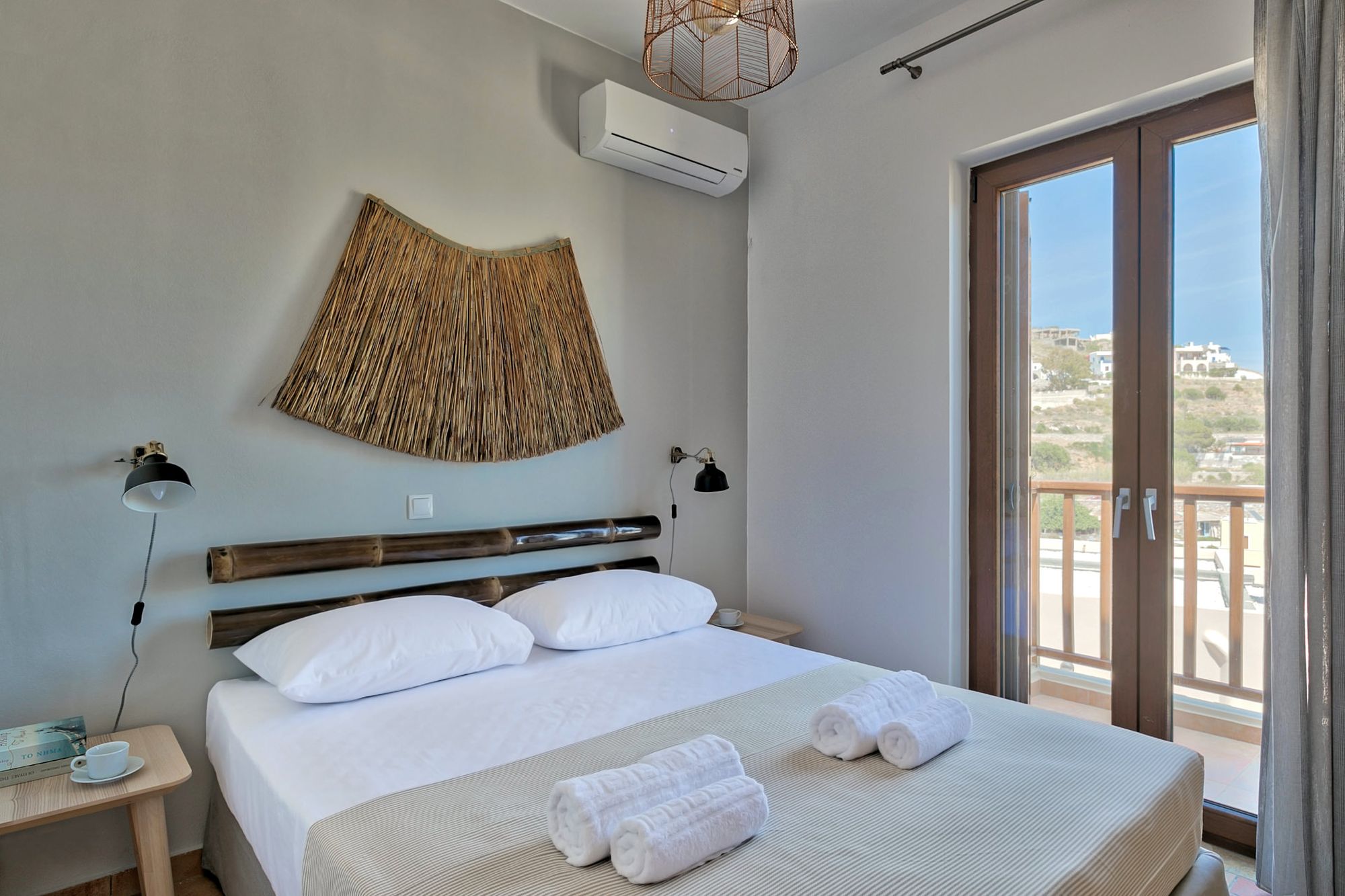 Air-conditioned bedroom with double bed, black wall lights, wooden bed side tables, a big wall wicker deco element and wooden bamboo masts over the bed.