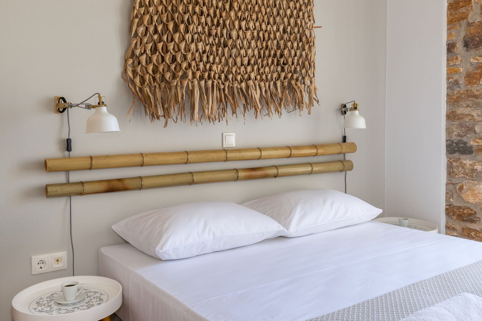 A double bed with white sheets in a stone-built bedroom, two white bedside tables, two gold-white wall lights and the wall over the bed is decorated with bamboo masts and a wicker decorative element. 