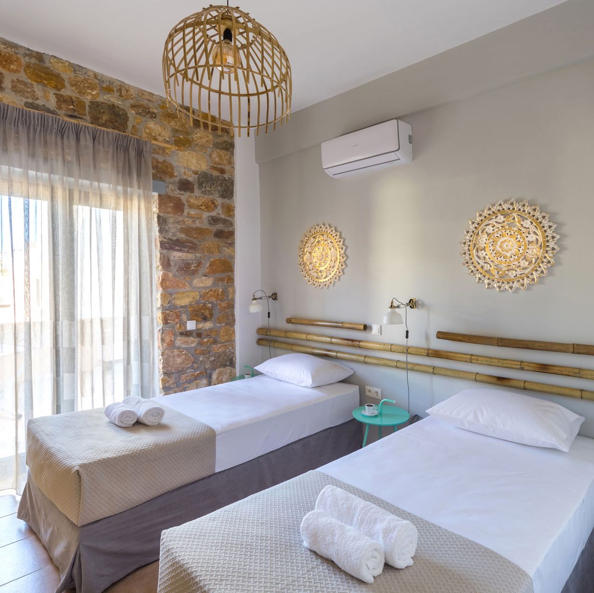 Twin bedroom of a stone built Syra Suite residence, blue bedside tables, white-gold wall lights, decorative masts and round gold metallic decorative elements on the wall over the beds.