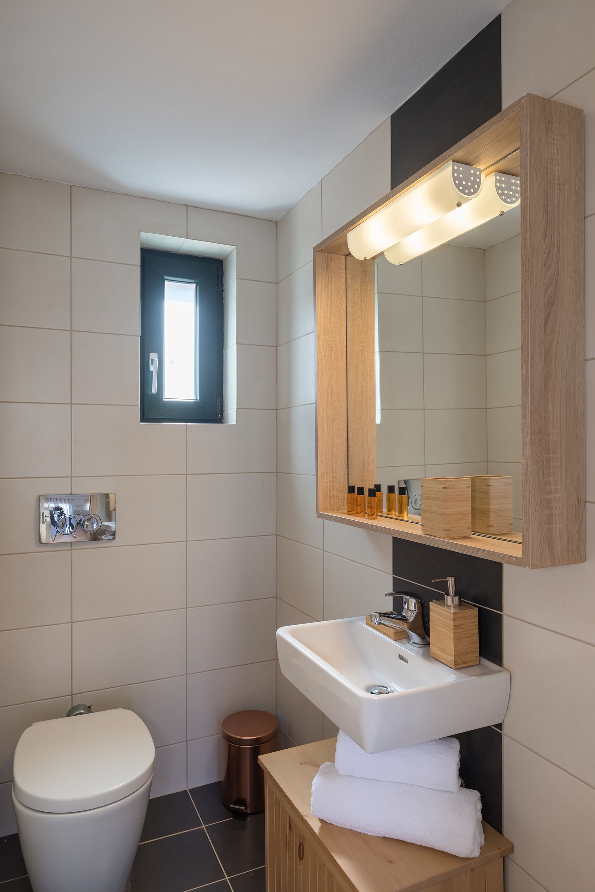 Bathroom with white and gray tiles, a white washbasin and a big wooden mirror.