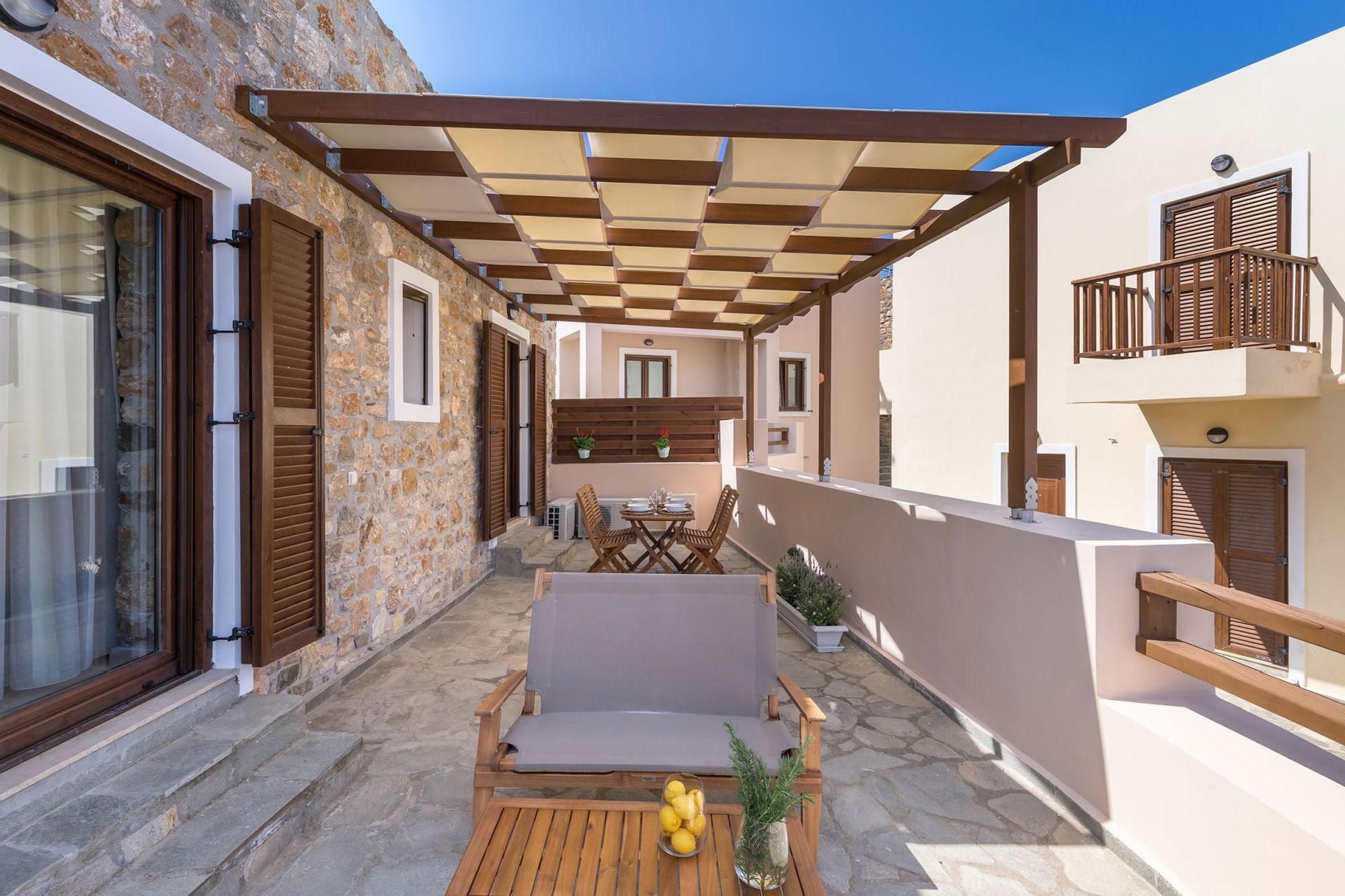 Veranda of a stone-built residence with pergola, furnished with a dining table and a lounge.