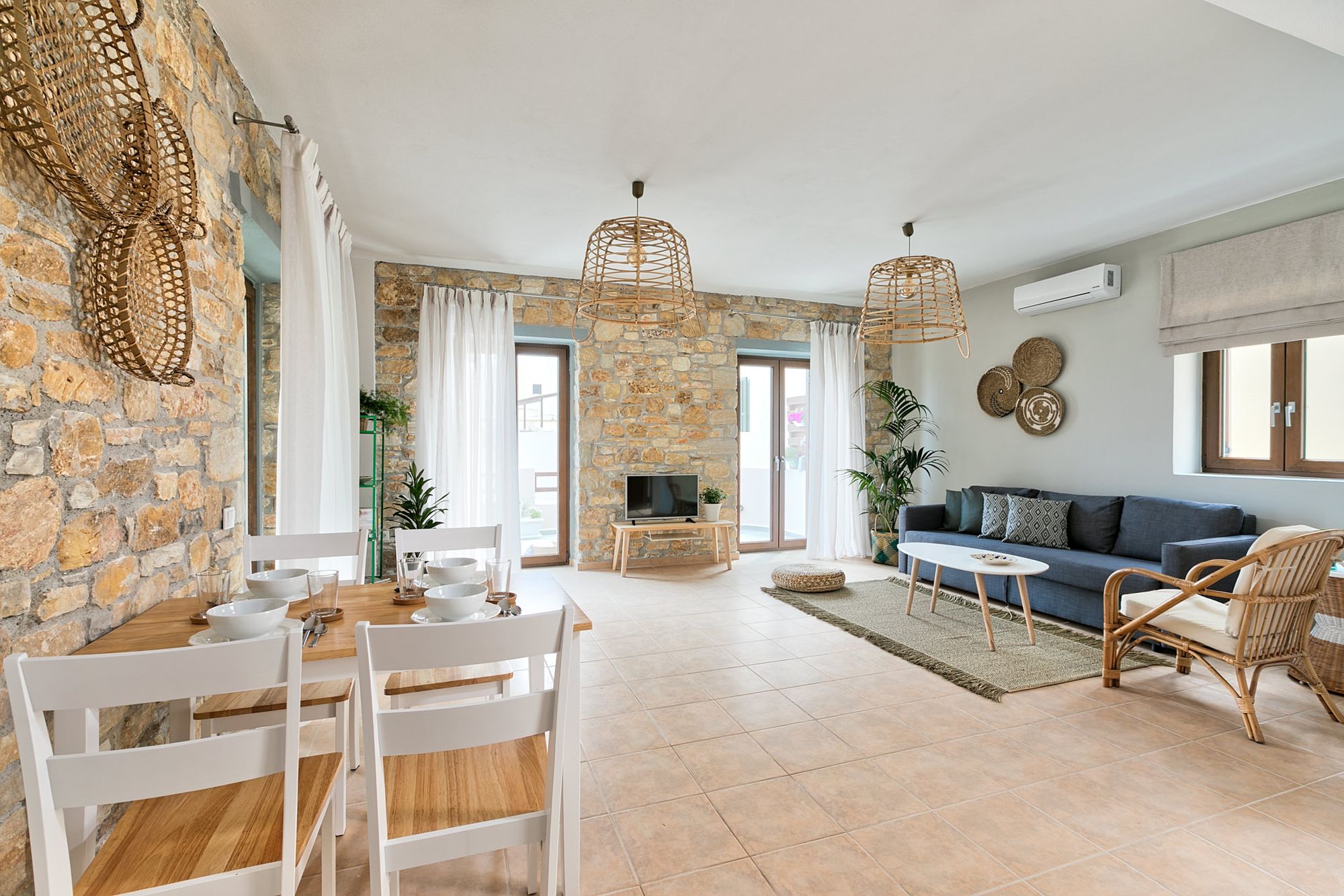 Very spacious living room of a stone-built residence with a blue sofa, a white wooden coffee table, an armchair, a dining table and wall wicker decorative elements.