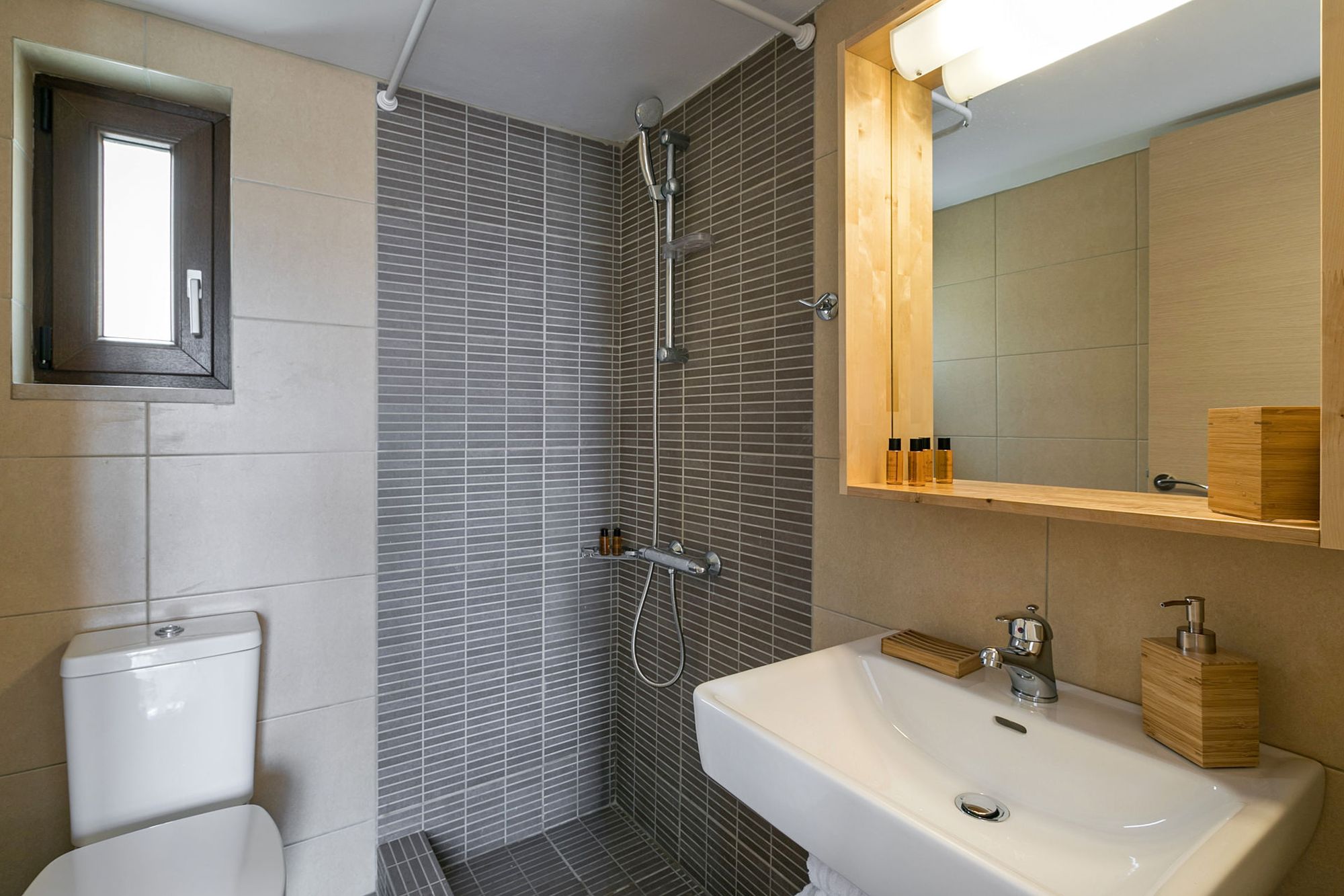 Modern bathroom with shower, big white washbasin, big wooden mirror and tiles in grey and beige tones.