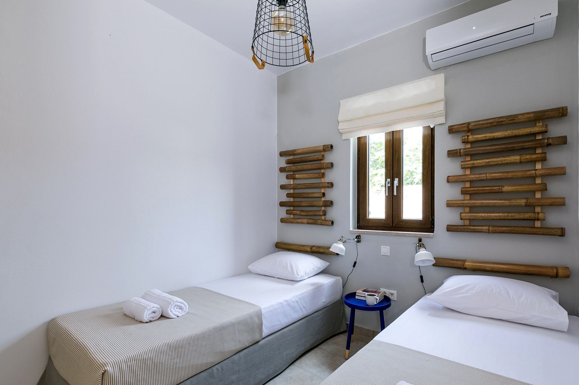 Modern twin bedroom with brown bamboo masts over the beds, a metallic blue bedside table, white wall lights and a window between the beds.