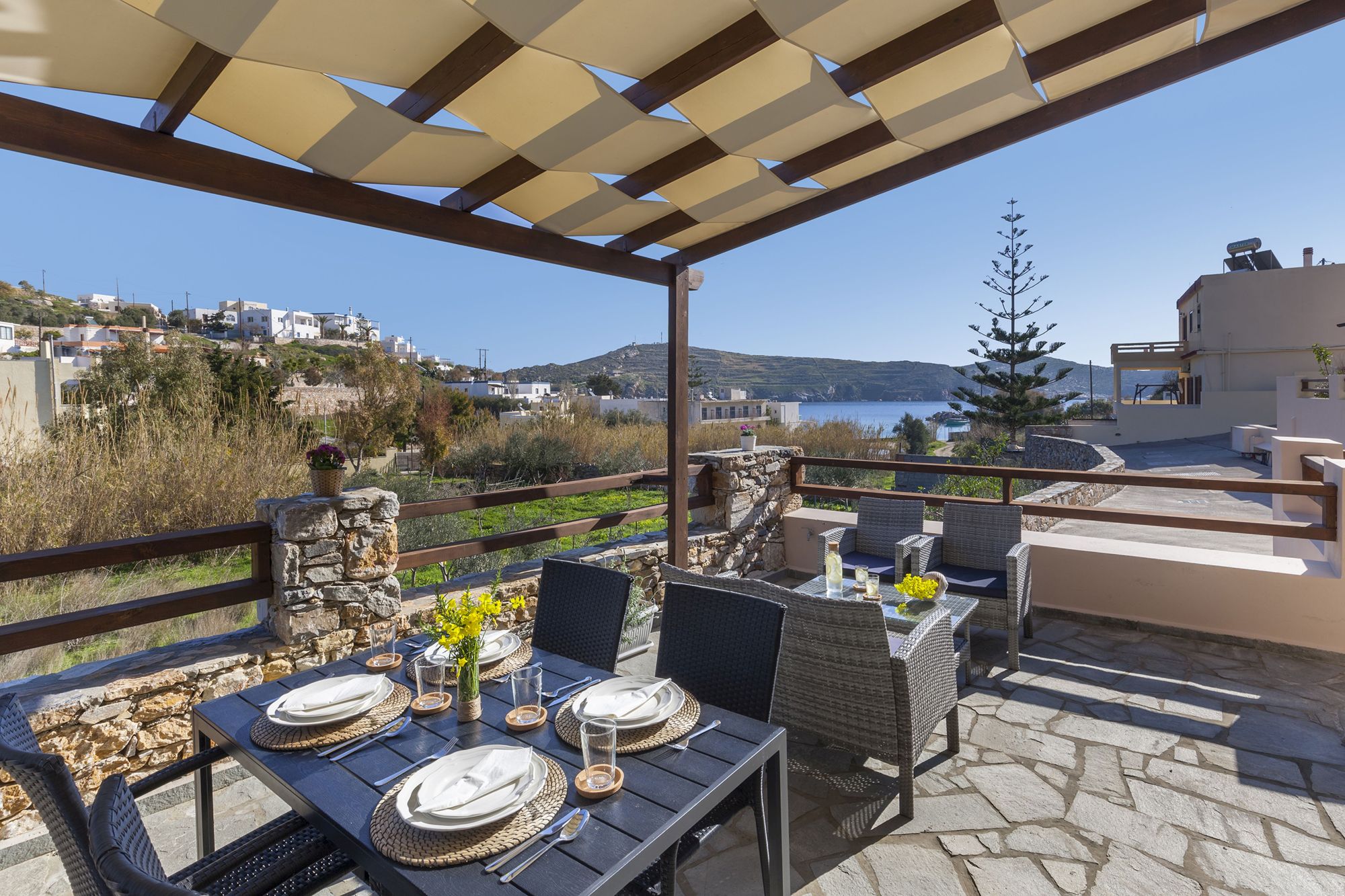 Sea view veranda of a stone-built residence with pergola, lounge and dining table with white dinnerware on it.