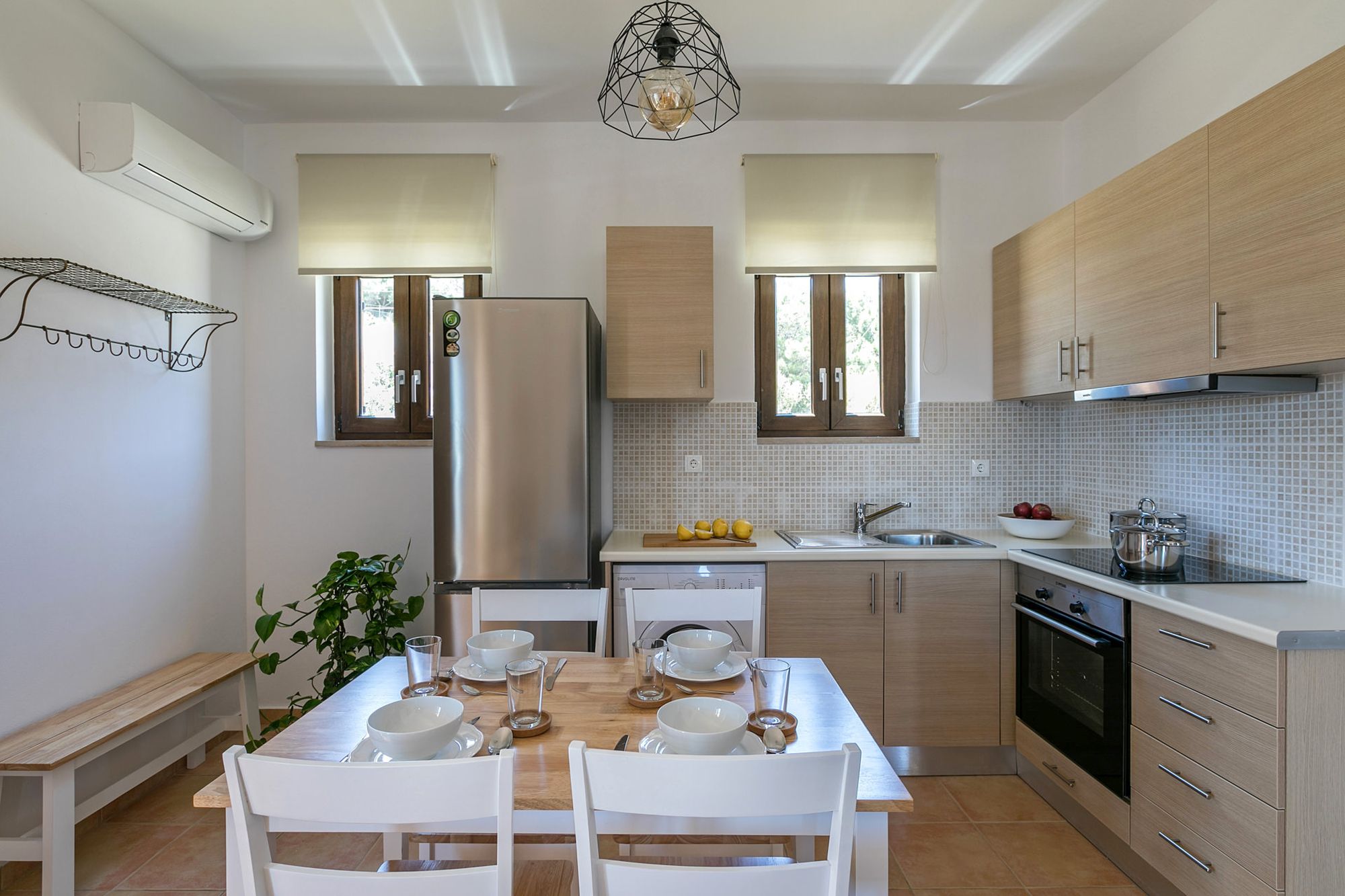 Syra Suites modern kitchen with beige cabinets, fully equipped with a big inox fridge, an electric cooker oven, a wooden bench and wooden dining table with four white chairs.