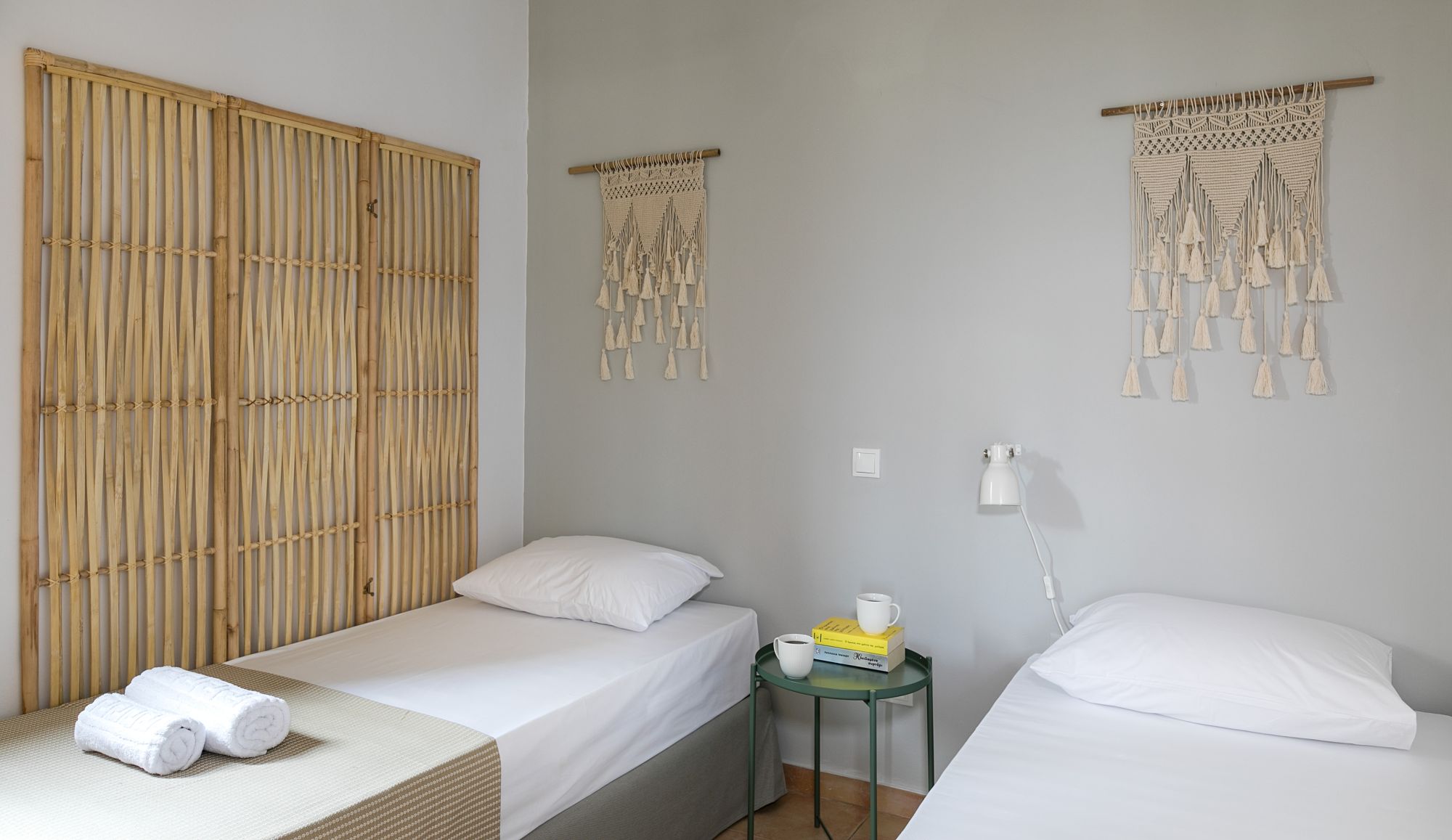 Twin bedroom with a metallic green bedside table between the two beds, white wall lights and boho decoration with a wall macramé and a decorative wicker screen.