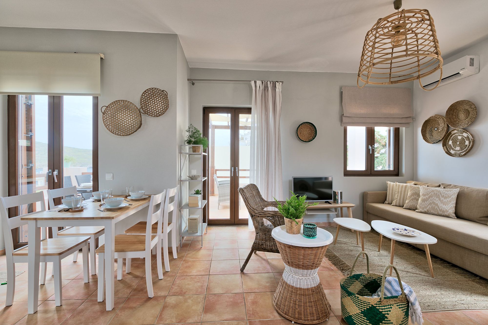 Living room with a big beige sofa, two modern white coffee tables in different sizes, a flat-screen TV, flowerpots, a metallic white bookcase and a white dining table.