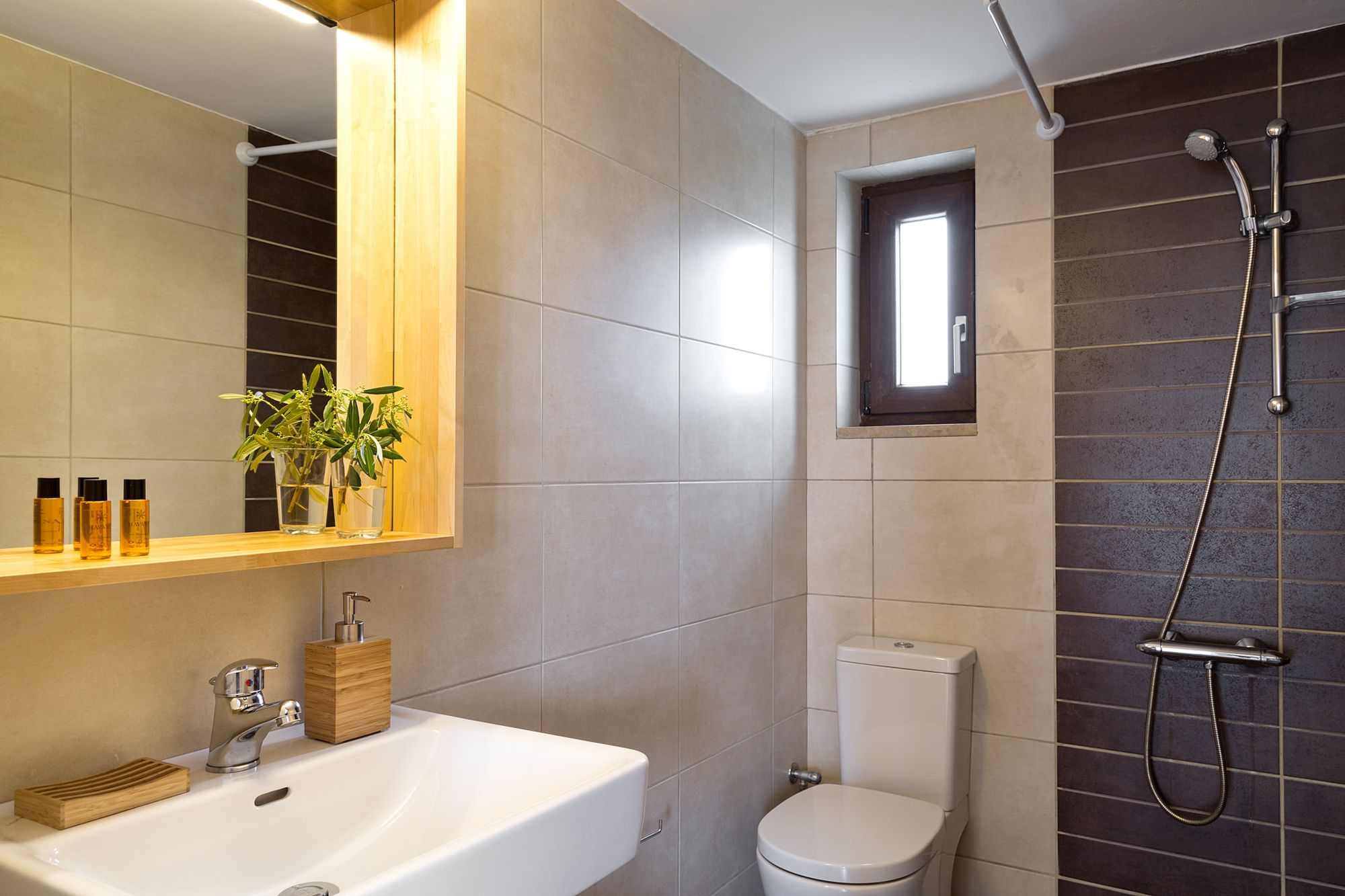 Μοντέρνο μπάνιο με ντουζιέρα, ένα μεγάλο λευκό νιπτήρα, ένα ξύλινο καθρέφτη και πλακάκια σε μπεζ και γκρι χρώματα.