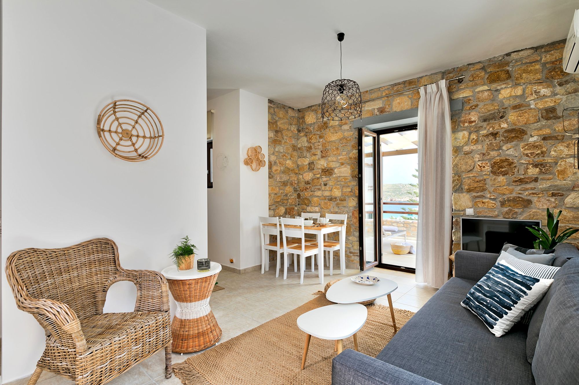 The living room of a stone built Syra Suite residence with a gray sofa, an armchair, two white coffee tables in different sizes, a flat screen TV and a dining table with four white chairs.