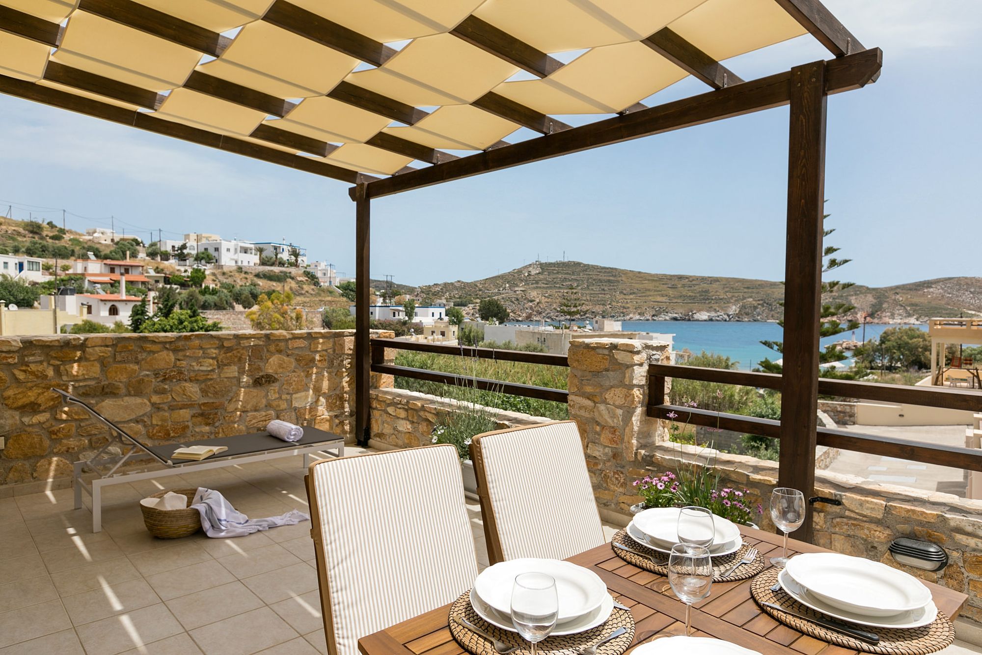 A sea view stone-built veranda with pergola, furnished with a wooden dining table and one sunbed.