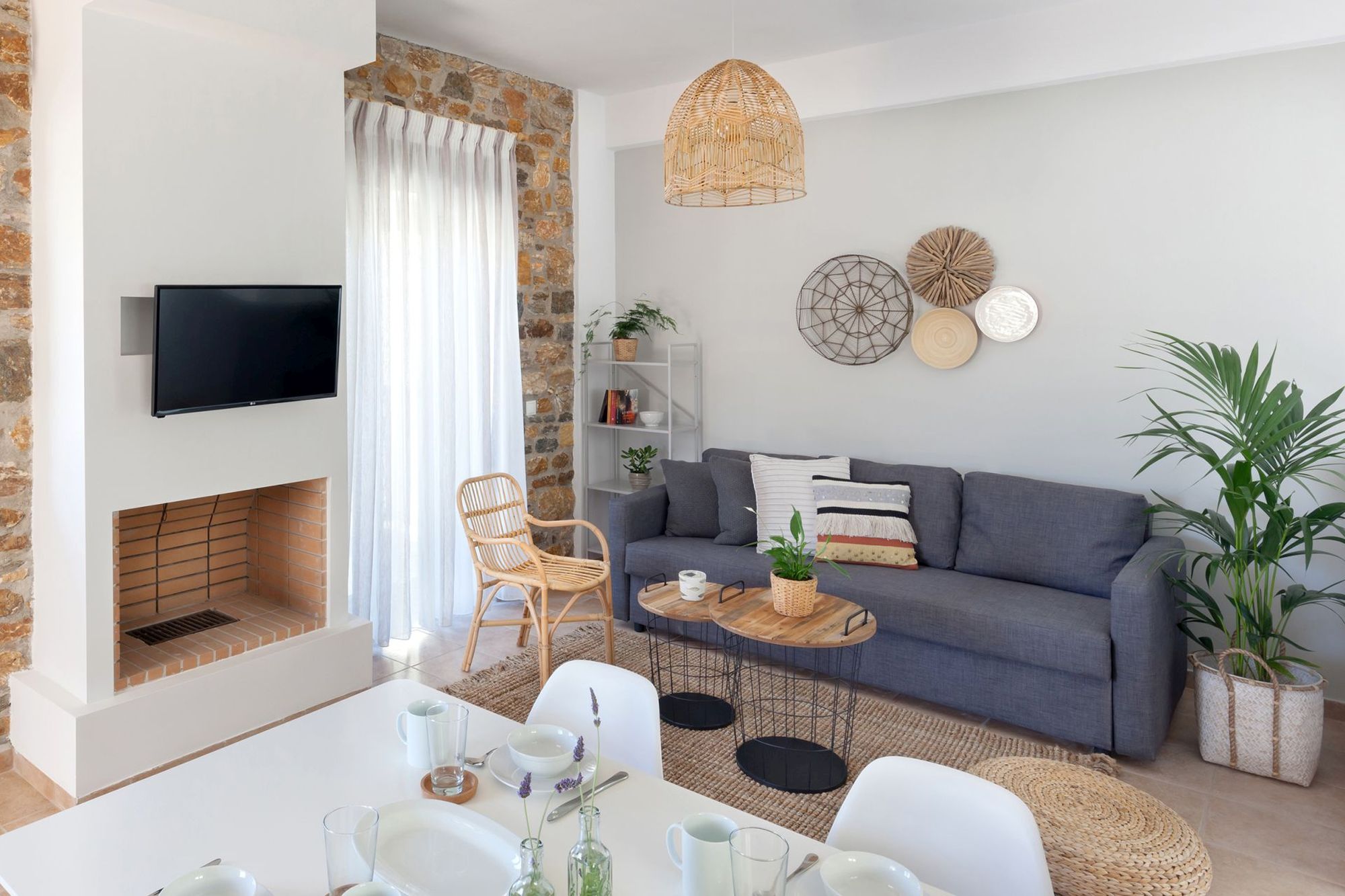 The living room of a stone built Syra Suite residence with fireplace, flat screen TV, big gray sofa, two metallic coffee tables in different sizes, an armchair and a dining table.