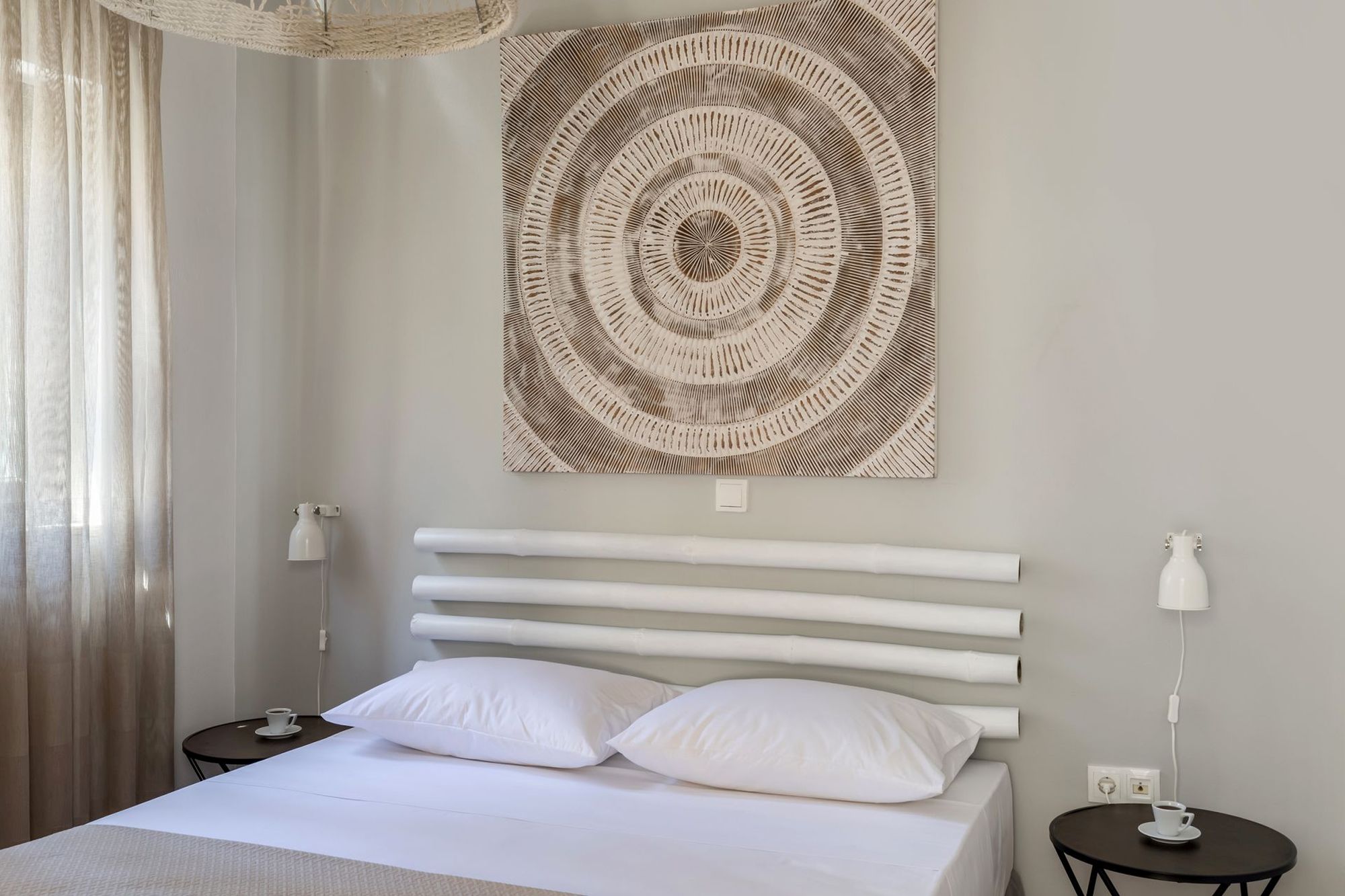 Bedroom with a double bed, metallic black bedside tables, wall white lights, decorative white bamboo masts and a big square decorative element in earth tones on the wall over the bed.