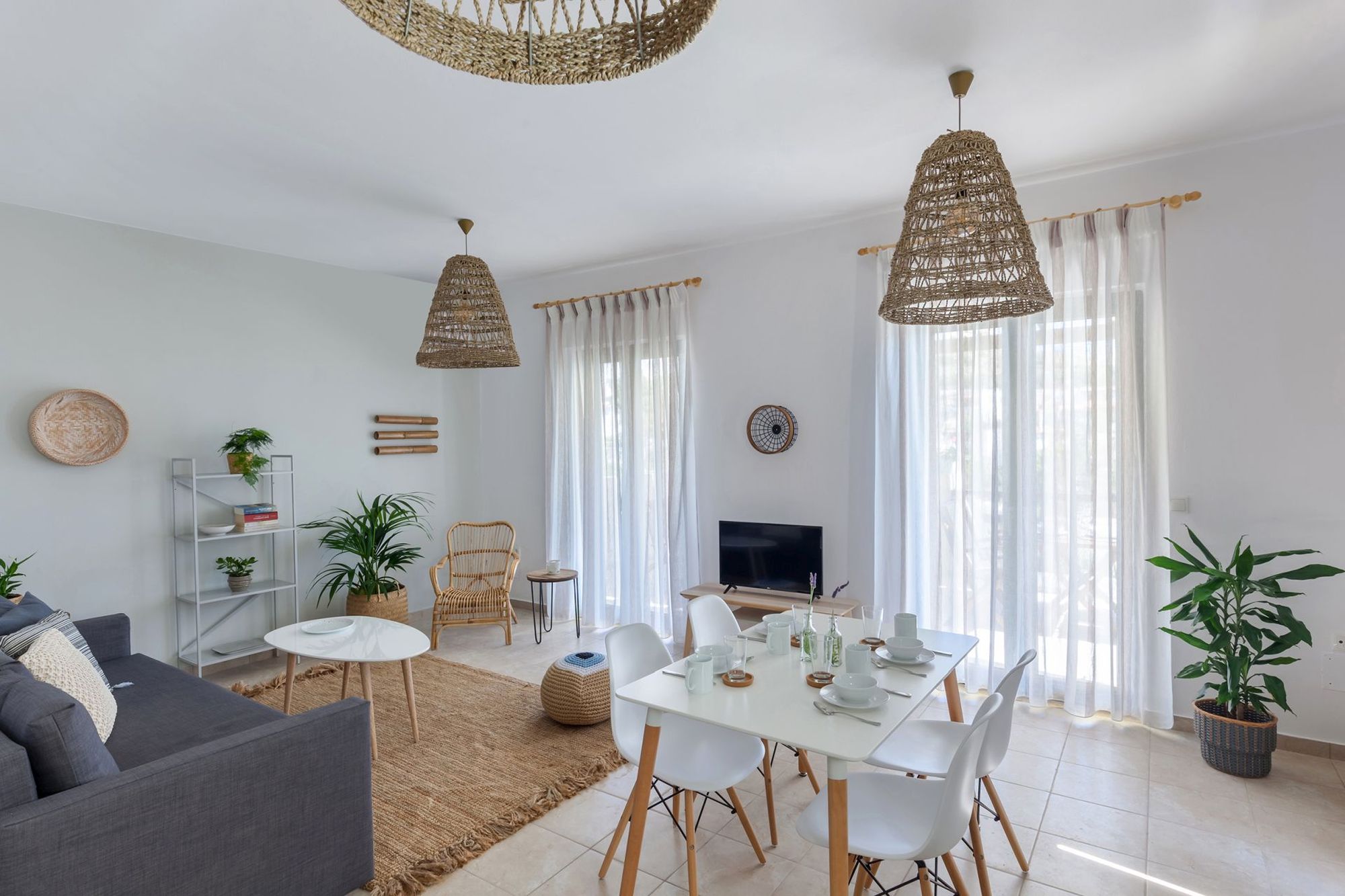 Spacious living room with gray sofa, white coffee table, metallic bookcase, wooden-white dining table and boho decoration with wicker ceiling lights and deco elements.