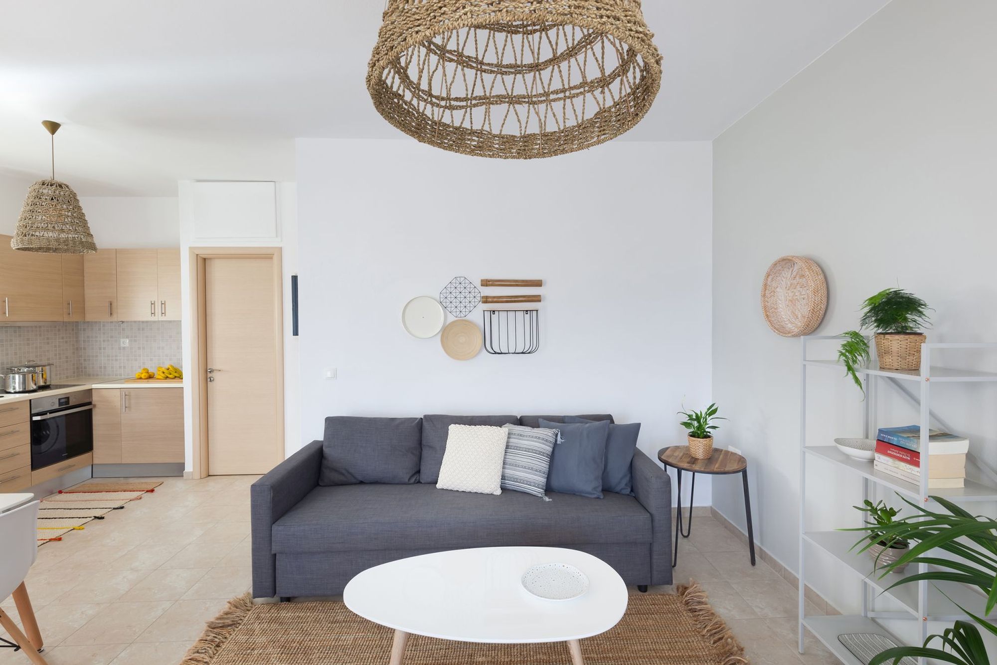 Living room with gray sofa, modern coffee table, white metallic bookcase and the kitchen in the background.