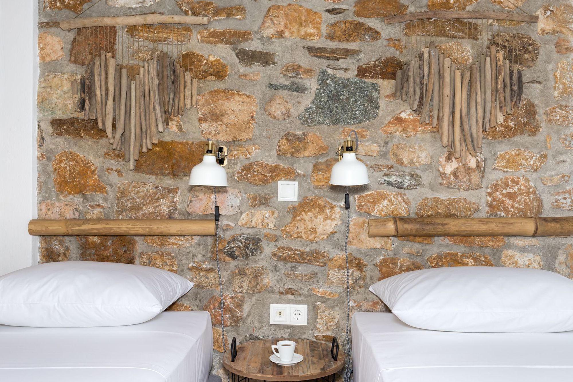 Twin bedroom of a stone built Syra Suite residence with a wooden bedside table between the two beds, white wall lights and boho decoration with bamboo masts on the wall Over the beds.