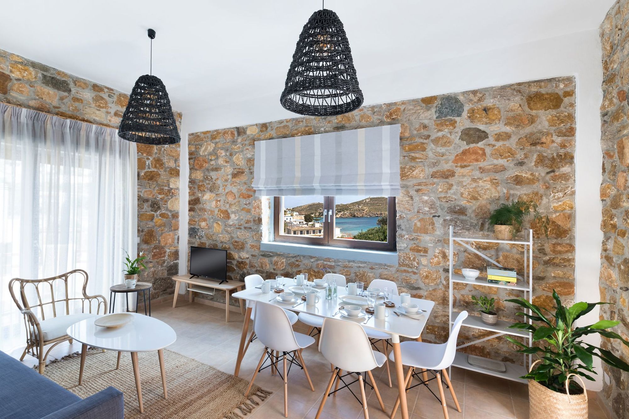 Dining area in same space with the living room of a stone built Syra Suite residence with a white dining table for six persons and a sea view window over the table.
