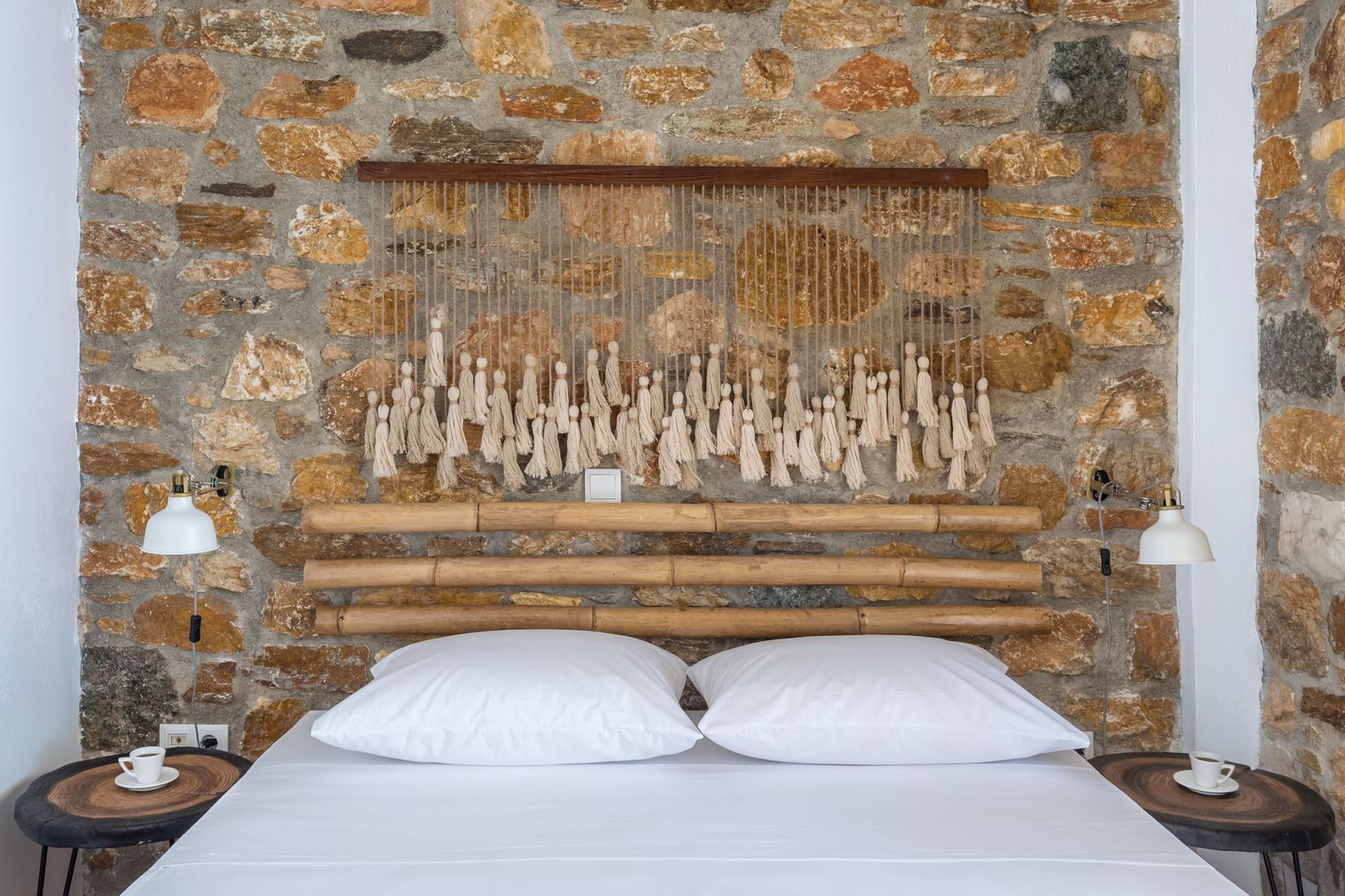 A stone-built bedroom with a double bed, two wooden bedside tables, white wall lamps beautifully decorated in boho style with a macramé over the bed.